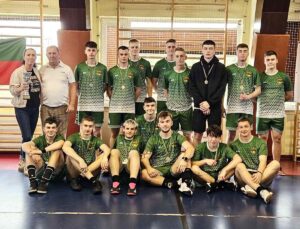 Szkolna drużyna sportowa w strojach koloru zielonego i białego oraz jeden zawodnik w czarnym stroju w hali sportowej. W tle drabinki sportowe. Dwie osoby dorosłe. Jedna z nich trzyma trofeum sportowe. Zawodnicy mają na szyjach medale. 
