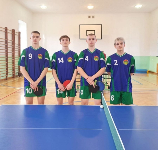 4 zawodników w hali sportowej przy stole do tenisa stołowego, stojących w strojach sportowych koloru niebieskiego, zielonego i białego z numerami startowymi na koszulkach i spodenkach. Trzech z nich trzyma również paletki tenisowe.