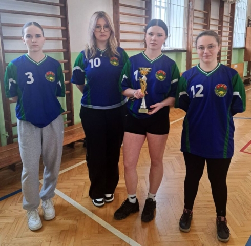 4 zawodniczki w strojach sportowych, stojące w hali sportowej, w tle drabinki gimnastyczne. Jedna z nich trzyma w ręku trofeum.