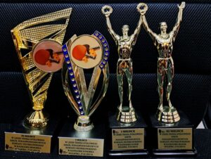 4 trofea sportowe, 2 w kształcie sportowców unoszących ręce w geście zwycięstwa, dwa w formach pucharów z symbolami rakietek tenisowych i piłek. Na dole napisy wielkimi literami I / III miejsce w Lidze tenisa stołowego szkół ponadpodstawowych w kategorii junior / juniorka (I miejsce) , senior / seniorka (III miejsce), Wolbórz 27.03.2025r. 