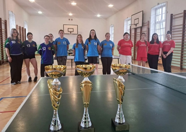 Sala gimnastyczna, w tle kosze do koszykówki oraz drabinki sportowe. 3 drużyny zawodniczek w strojach sportowych, po 4 osoby w każdej drużynie. Na pierwszym planie stół do tenisa stołowego z 3 pucharami.