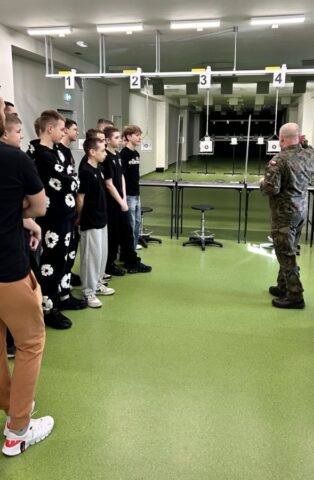 mks_piotrkowianin_strzelnica01 Grup młodzieży stoi w pomieszczeniu strzelnicy i uczestniczy w szkoleniu prowadzonym przez Instruktora. W tle stanowiska strzelnicze oraz tarcze.