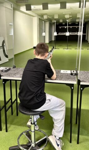 mks_piotrkowianin_strzelnica02 Nastolatek siedzący na stanowisku strzelniczym obiera cel. W ręku trzyma broń, z której będzie strzelał.