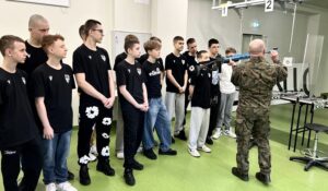 Grupa młodzieży stoi w pomieszczeniu strzelnicy i obserwuje instruktora udzielającego wskazówek. 