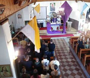 Wnętrze kościoła. W ławkach uczniowie i nauczyciele. Przy ołtarzy ksiądz odprawiający mszę wraz z ministrantami. Kościół przyozdobiony flagami koloru żółtego, żółto - białego i niebiesko - białego. Przy ołtarzu Krzyż zasłonięty fioletowym suknem postnym. 