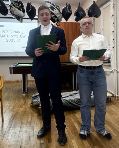 Dwóch uczniów w dużej klasie, trzymają w ręku teczki prowadzą uroczystość pożegnania maturzystów w internacie. W tle ekran z wyświetloną prezentacją, tekst Pożegnanie maturzystów 2025, przy suficie zawieszone czarne i czarno białe balony- te ostatnie z napisem Congrats You did it! Graduate. balony