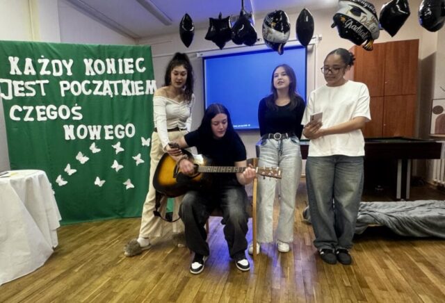 Uroczystość pożegnania maturzystów w internacie. Na pierwszym planie 4 uczennice, jedna gra na gitarze, wszystkie śpiewają. W tle stolik przykryty białym obrusem, tablica osłonięta zielonym materiałem z białym napisem "Każdy koniec jest początkiem czegoś nowego" i białymi dekoracjami w kształcie motyli. Przy suficie zawieszone balony czarne oraz czarno- białe z napisem Congrats! you did it! Graduate.