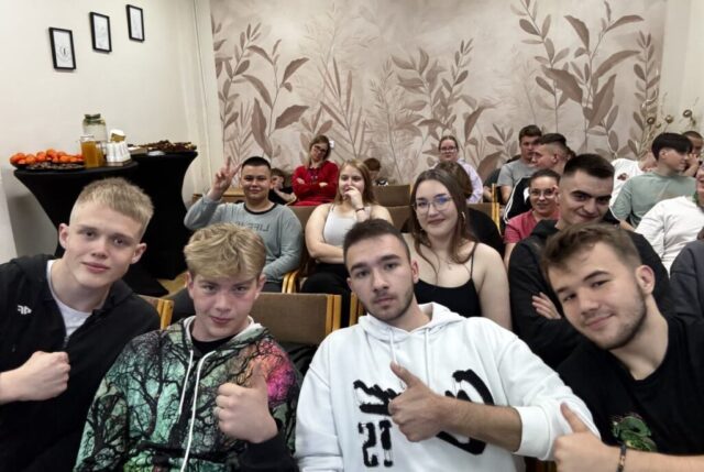 Grupa uczniów wraz z opiekunem, siedzą w dużej klasie. W tle ściana z fototapetą z motywem roślinnym. Po lewej stornie czarne okrągłe stoliki z poczęstunkiem, owocami, napojami i ciastami.
