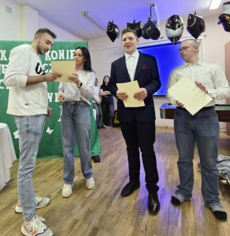 Uczeń odbiera z rąk opiekuna dyplom. Po prawej stronie dwóch uczniów, prowadzących uroczystość. W tle tablica przykryta zielonym materiałem z częściowo widocznym napisem koloru białego "Każdy koniec jest początkiem czegoś nowego...". Na drugim planie dwie uczennice- członkinie zespołu wokalnego. U góry przy suficie zawieszone balony czarne i czarno-białe z napisem Congrats. Yyou did it! Graduate.