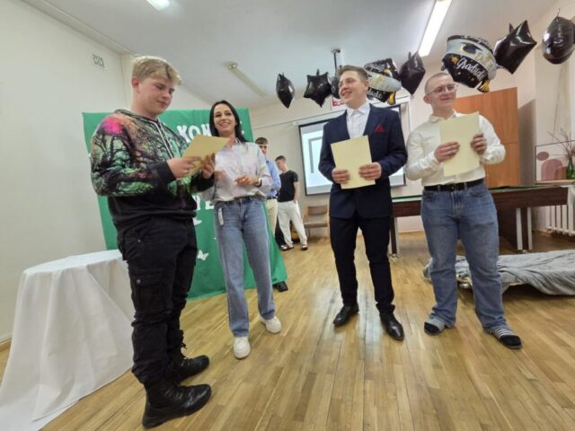 Uczeń odbiera z rąk opiekuna dyplom. Po prawej stronie dwóch uczniów, prowadzących uroczystość. W tle tablica przykryta zielonym materiałem z częściowo widocznym napisem koloru białego "Każdy koniec jest początkiem czegoś nowego...". Na drugim planie dwóch innych uczniów. U góry przy suficie zawieszone balony czarne i czarno-białe z napisem Congrats. Yyou did it! Graduate.