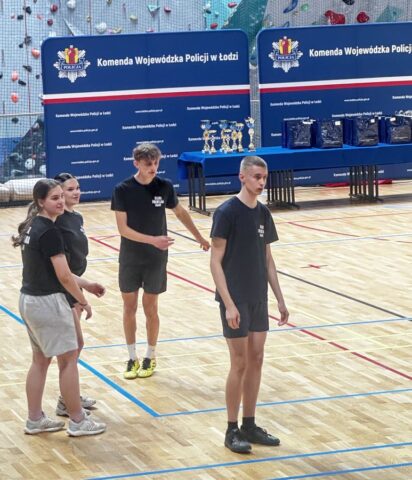 puchar_komendanta_policji05 4 zawodników w strojach sportowych w hali sportowej. W tle granatowe banery reklamowe Komendy Wojewódzkiej Policji w Łodzi, ławki przykryte granatowym suknem, na których stoją nagrody i puchary.