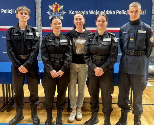 puchar_komendanta_policji10 4 uczniów w umundurowaniu klasy policyjnej wraz z opiekunem w hali sportowej na tle baneru reklamowego Komendy Wojewódzkiej Policji w Łodzi.