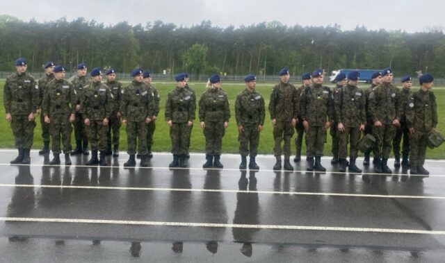 Grupa uczniów w umundurowaniu klas wojskowych stoi na zewnątrz. W tle drzewa i teren sportowy jednostki wojskowej.