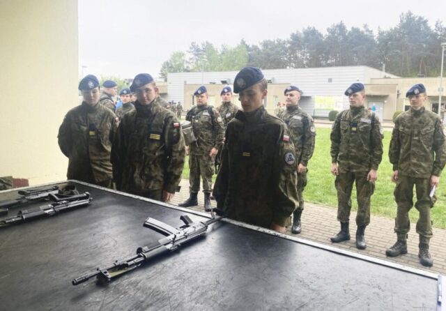 Młodzież w umundurowaniu klas wojskowych stoi przed stołem, na którym leży broń. W tle zabudowania oraz inni uczestnicy Spartakiady.