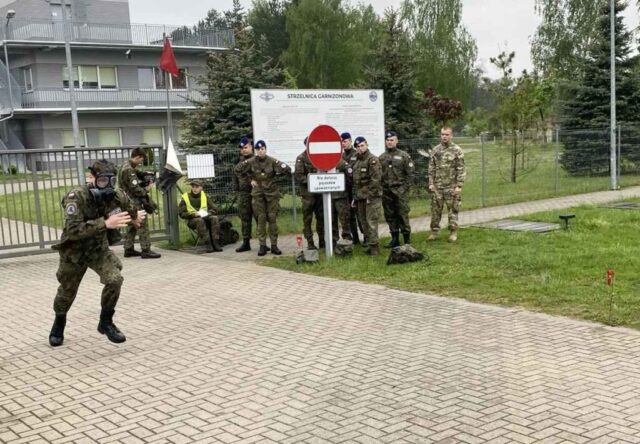 Młodzież w umundurowaniu klas wojskowych na zewnątrz. Jeden zawodnik w masce p. gazowej biegnie. W tle budynek o raz ogrodzony teren. Widoczny także masz z czerwoną flagą oraz tablica informacyjna z nazwą i logo Strzelnica Garnizonowa.
