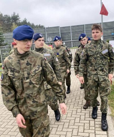 Grupa młodzieży w umundurowaniu klas wojskowych idzie dwójkami. W tle ogrodzony teren ekrany dźwiękoszczelne.
