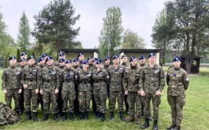 23 uczniów w umundurowaniu klas wojskowych stoi na zewnątrz w rzędach. W tle wiaty, altana i drzewa. 