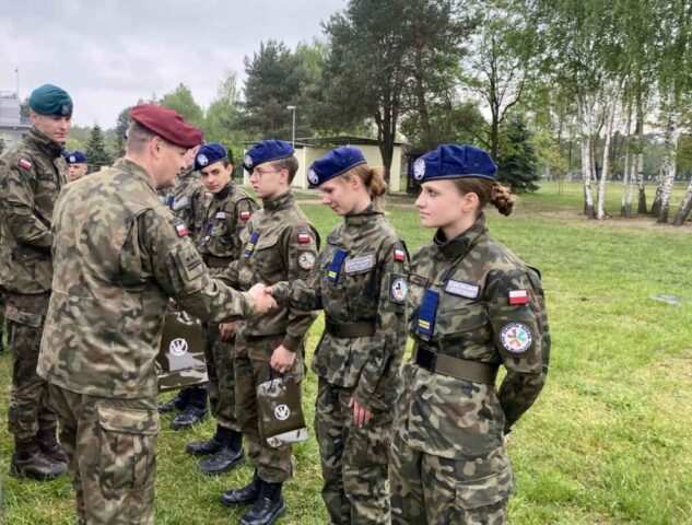Dwóch żołnierzy przekazuje młodzieży gratulacje i torby upominkowe w kolorze khakim. W tle wiaty i drzewa.