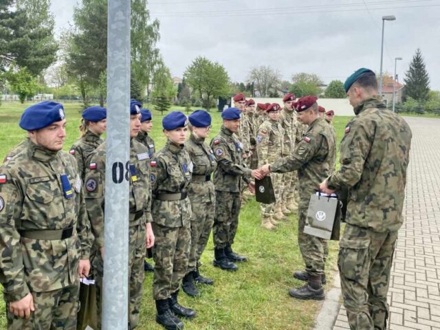 Dwóch żołnierzy przekazuje młodzieży gratulacje i torby upominkowe w kolorze khakim. Widoczna także inna drużyna biorąca udział w Spartakiadzie. W tle zabudowania i drzewa.