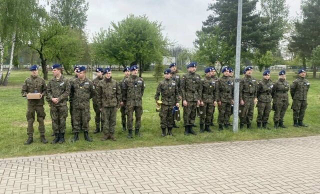 Młodzież ZS CKZ Bujny biorąca udział w Spartakiadzie stoi na zewnątrz. Wszyscy w umundurowaniu klas wojskowych i nakryciach głowy (granatowe berety). Część kadetów trzyma w rękach torby upominkowe, jeden ma też puchar i dyplom a kadet stojący po lewej strony trzyma też tekturowe pudełko. W tle ogrodzony teren oraz drzewa. .