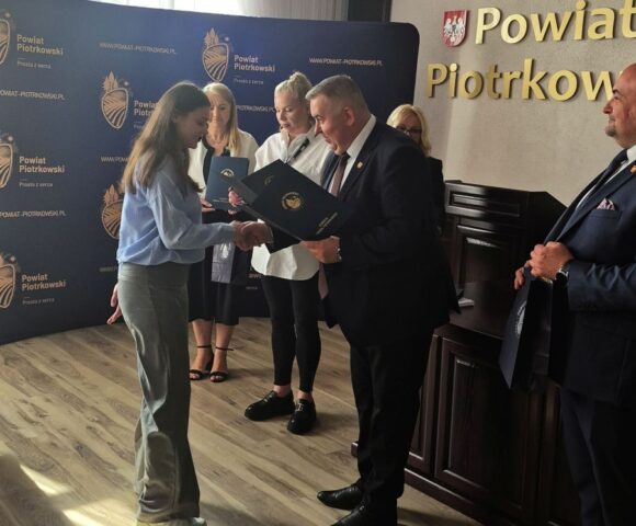 Uczennica odbiera z rąk Starosty dyplom. W tle p. Dyrektor ZS CKZ Bujny oraz przedstawiciele Starostwa Powiatowego. Widoczny również baner reklamowy starostwa z logo.