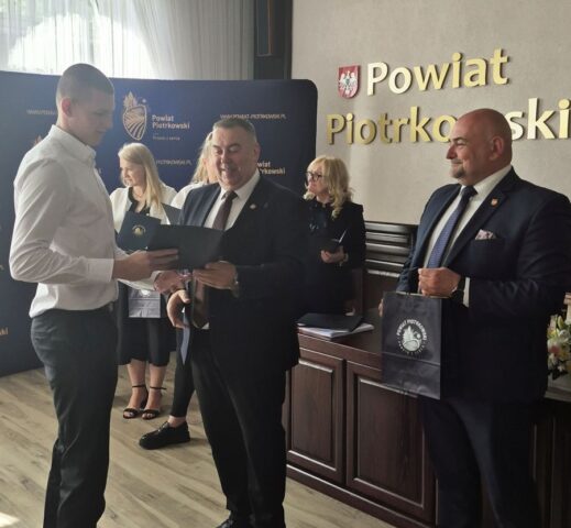 Uczeń odbiera z rąk p. Starosty dyplom. W tle p. Dyrektor ZS CKZ Bujny oraz przedstawiciele Starostwa Powiatowego oraz po prawej stronie p. Wicestarosta trzymający torbę reklamową Starostwa powiatowego. . Widoczny również baner reklamowy starostwa z logo.