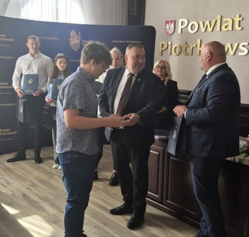 Uczeń odbiera z rąk p. Starosty dyplom. W tle inni uczniowie ZS CKZ Bujny, stypendyści oraz przedstawiciele Starostwa Powiatowego oraz po prawej stronie p. Wicestarosta trzymający torbę reklamową Starostwa powiatowego. Widoczny również baner reklamowy starostwa z logo.