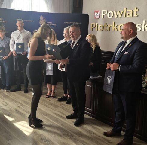 Uczennica odbiera dyplom z rąk p. Starosty. Po prawej stronie p. wicestarosta, trzyma w ręku torbe reklamową z logo Powiatu. W tle przedstawiciele Starostwa, p. dyrektor ZS CKZ Bujny i inni uczniowie- stypendyści. Widoczny też baner reklamowy powiatu piotrkowskiego z logo i adresem strony internetowej.