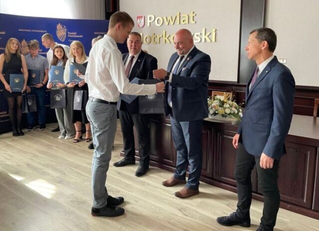 Uczeń odbiera torbę reklamową z rąk p. Wicestarosty. Obok stoi p. Starosta. W tle p. dyrektor ZS CKZ Bujny oraz inni uczniowie, stypendyści. Widoczny też baner reklamowy Starostwa z logo.