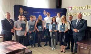 Grupa uczniów, stypendystów z p. Dyrektor oraz przedstawicielami Starostwa Powiatowego w pomieszczeniu. W tle baner reklamowy i logo Powiatu Piotrkowskiego. 