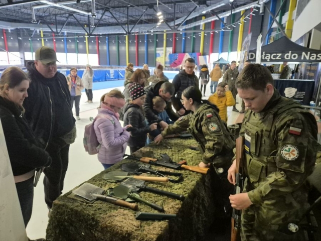 Duża hala sportowa. Stoisko promocyjne Szkoły i klasy OPW. Na stole przykrytym siatką maskującą zaprezentowana broń oraz inny sprzęt wojskowy. Na stoisku uczniowie w umundurowaniu z flagą Polski na ramieniu i logo szkoły, objaśniają zwiedzającym oglądany sprzęt. W tle baner z napisem Dołącz do Nas.