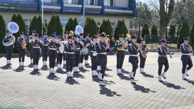 Grupa muzykantów z orkiestry. Wszyscy w mundurach koloru stalowego oraz nakryciach głowy i białych butach. Grają na roznych instrumentach muzycznych. Stoją na zewnątrz, w tle budynek.