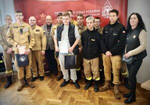 Grupa młodzieży i dorosłych stojąca w pomieszczeniu. W tle czerwony baner z częściowo widocznym napisem Państwowa Straż Pożarna, logo oraz godłem. Część młodzieży w umundurowaniu klasy strażackiej. Jeden dorosły również w mundurze strażackim, pozostałe dwie osoby dorosłe oraz jeden uczeń w strojach codziennych. Dwóch uczniów trzyma dyplomy oraz torby.