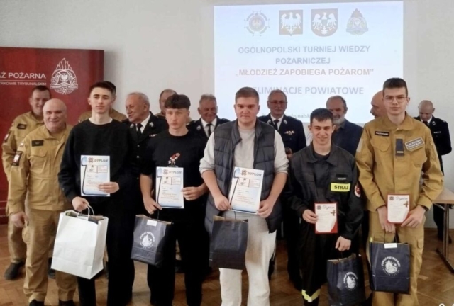 Grupa młodzieży szkolnej oraz dorosłych stojących w pomieszczeniu. W tle slajd z logo patronów oraz nazwą turnieju: Ogólnopolski Turniej Wiedzy Pożarniczej "Młodzież zapobiega pożarom" Eliminacje Powiatowe. Dorośli oraz część młodzieży w umundurowaniu. Stojący na pierwszym planie uczniowie trzymają w rękach dyplomy oraz torby z logo Powiatu Piotrkowskiego.