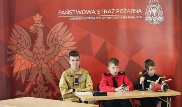 3 uczniów siedzących za stołem. W tle czerwony baner z napisem Państwowa Straż Pożarna Komenda Miejska PSP w Piotrkowie Trybunalskim. Po lewej stronie godło, po prawej logo PSP.