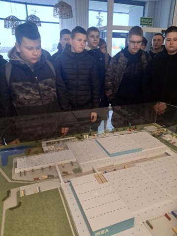 Grupa młodzieży w pomieszczeniu. Stoją nad makietą centrum logistycznego, zainteresowani oglądanym eksponatem.