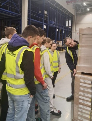 Grupa młodzieży w kamizelkach odblaskowych z napisem Visitors na plecach. Stoją w magazynie logistycznym i oglądają prezentowane przez przewodnika towary na paletach.