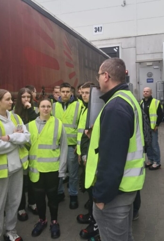 Grupa młodzieży w kamizelkach odblaskowych w hali logistycznej. Słuchają dwóch panów oprowadzających ich po obiekcie. W tle kontener.