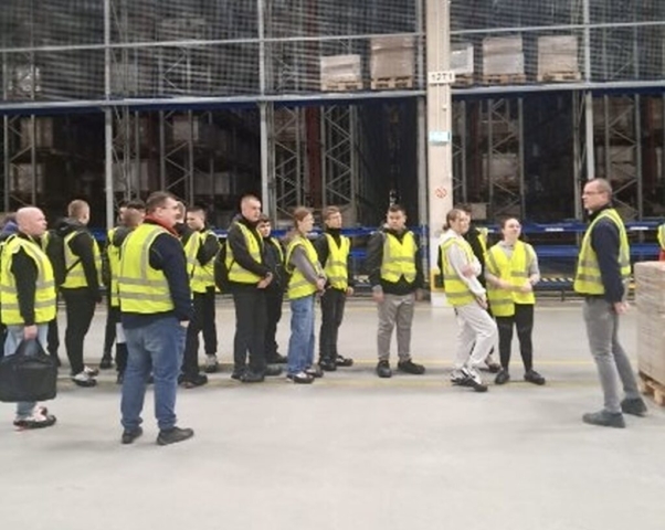 Grupa młodzieży i 3 przewodników w kamizelkach odblaskowych stoją w magazynie wysokiego składowania.