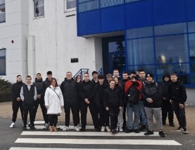 Grupa młodzieży wraz z opiekunem. Stoją na zewnątrz, przed biało granatowym budynkiem biurowym. l