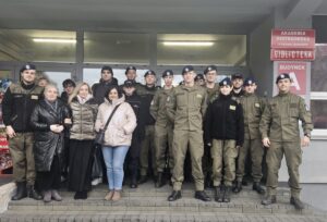 Grupa młodzieży wraz z opiekunami stoją przed budynkiem Akademii Piotrkowskiej. W tle wejście do uczelni oraz 3 czerwone tabliczki z napisami: Akademia Piotrkowska w Piotrkowie Trybunalskim, Biblioteka, Budynek A. Młodzież w większości w umundurowaniu klas wojskowych i policyjnych. 