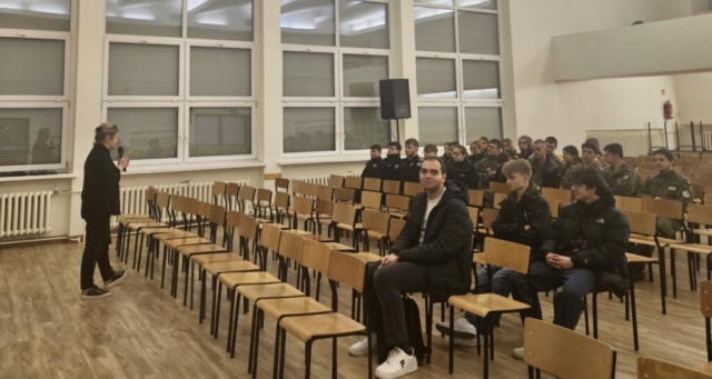 Młodzież siedzi w auli uczelni i słucha wystąpienia kobiety przemawiającej do nich przez mikrofon.