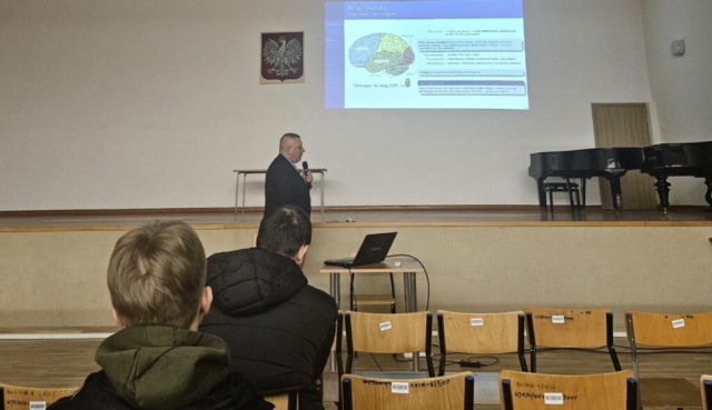 Aula uczelni, młodzież siedzi na krzesełkach i uczestniczy w prelekcji poświęconej zapamiętywaniu. W tle wyświetlana prezentacja - Mózg- budowa. Mężczyzna w garniturze trzyma mikrofon i prowadzi wykład. Na ścianie, po lewej stronie, widoczne godło Polski a na scenie, po prawej stronie fortepian.