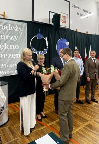 Absolwent technikum leśnego w mundurze wręcza bukiety kwiatów p. Dyrektor i wychowawcy. Widoczni także zaproszeni na uroczystość goście. W tle stojak z flagami Polski.