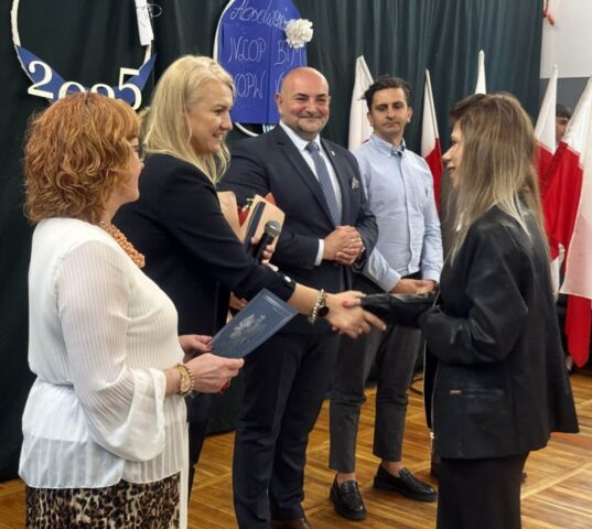 Uroczystość zakończenia roku szkolnego klas maturalnych. Przedstawicielka rady rodziców odbiera gratulacje z rąk p. Dyrektor. Towarzyszą p. Wicedyrektor, p. Wicestarosta i zaproszeni goście. Sala gimnastyczna udekorowana stosownie do okoliczności, w tle stojak z flagami Polski.