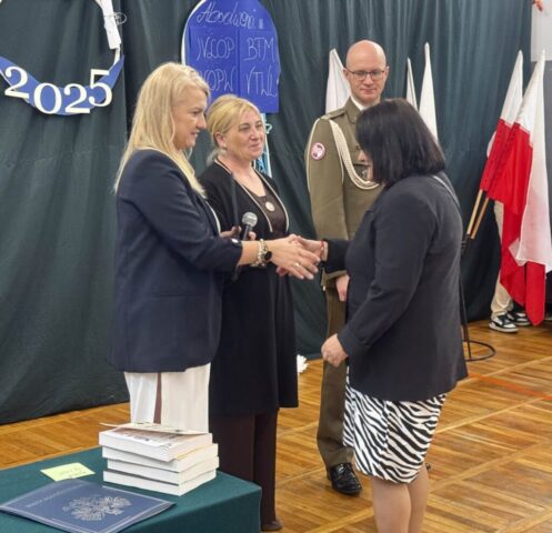 Kobieta odbiera z rąk p. Dyrektor gratulacje. Widoczny także wychowawczyni klasy oraz przedstawiciel służb mundurowych. Po lewej stronie stolik przykryty zielonym suknem, na którym widać nagrody książkowe oraz teczkę z orłem.