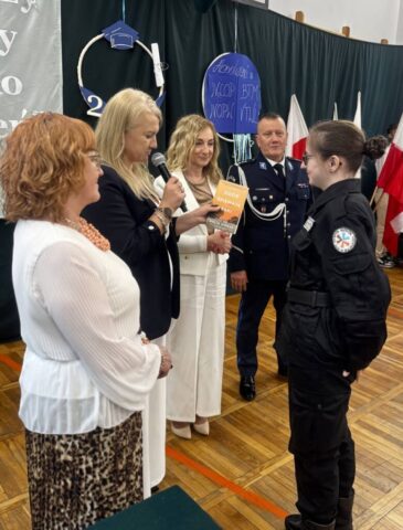 Absolwentka klasy policyjnej odbiera gratulacje i nagrodę książkowa z rąk p. Dyrektor. Towarzyszą p. Wicedyrektor, p. Wychowawczyni i przedstawiciel służb mundurowych policji. W tle stojak z flagami polski.