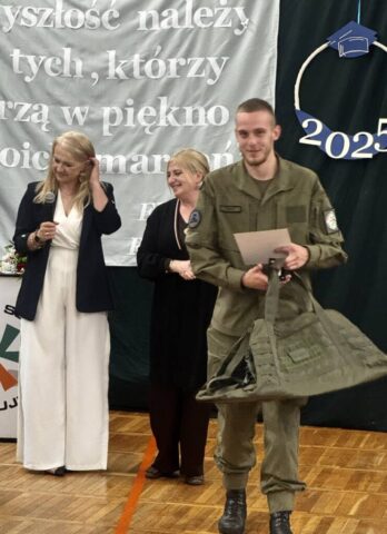 Uczeń klasy OPW w umundurowaniu trzymający w ręku nagrodę rzeczową i dyplom. W tle p. Dyrektor i p. wychowawczyni.
