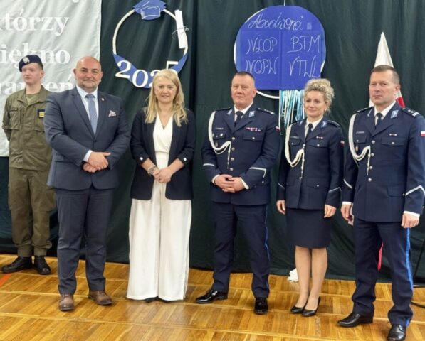 p. Dyrektor, p. Wicestarosta i przedstawiciele policji oraz uczeń klasy OPW na sali gimnastycznej podczas uroczystości zakończenia roku klas maturalnych. W tle hasło przewodnie uroczystości i dekoracje.