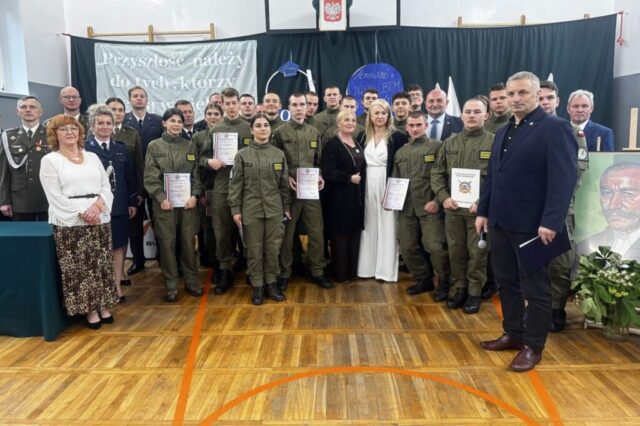 Uroczystość zakończenia roku szkolnego klas maturalnych. Absolwenci klasy OPW, Dyrekcja, wychowawca, koordynator klas OPW, zaproszeni goście, przedstawiciele służb mundurowych i władz lokalnych w udekorowanej na okoliczność sali gimnastycznej. Absolwenci trzymają w rękach certyfikaty i dyplomy.